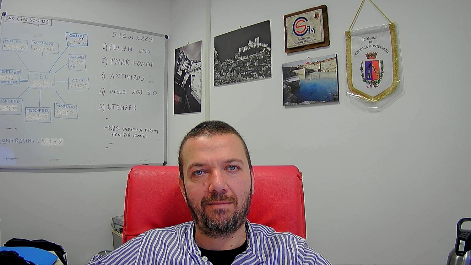 Alessandro Rossi, tecnico informatico dell’Ente, è seduto alla scrivania del suo ufficio. Indossa una camicia bianca a righe grigie.